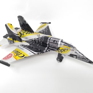 May include: A model airplane made from recycled aluminum cans. The plane is painted black with yellow accents and has the words "Schweppes" and "Canada Dry" printed on the wings.