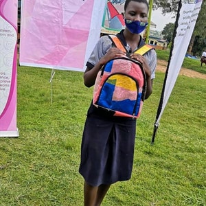 Pu&ograve; includere: Una giovane persona con una maschera facciale tiene uno zaino colorato con motivi multipli. Lo zaino ha una cerniera e spalline nere. La persona &egrave; in piedi su un prato erboso. Un cartello dietro la persona recita "SMART BAGS "KEEPING GIRLS IN SCHOOL."