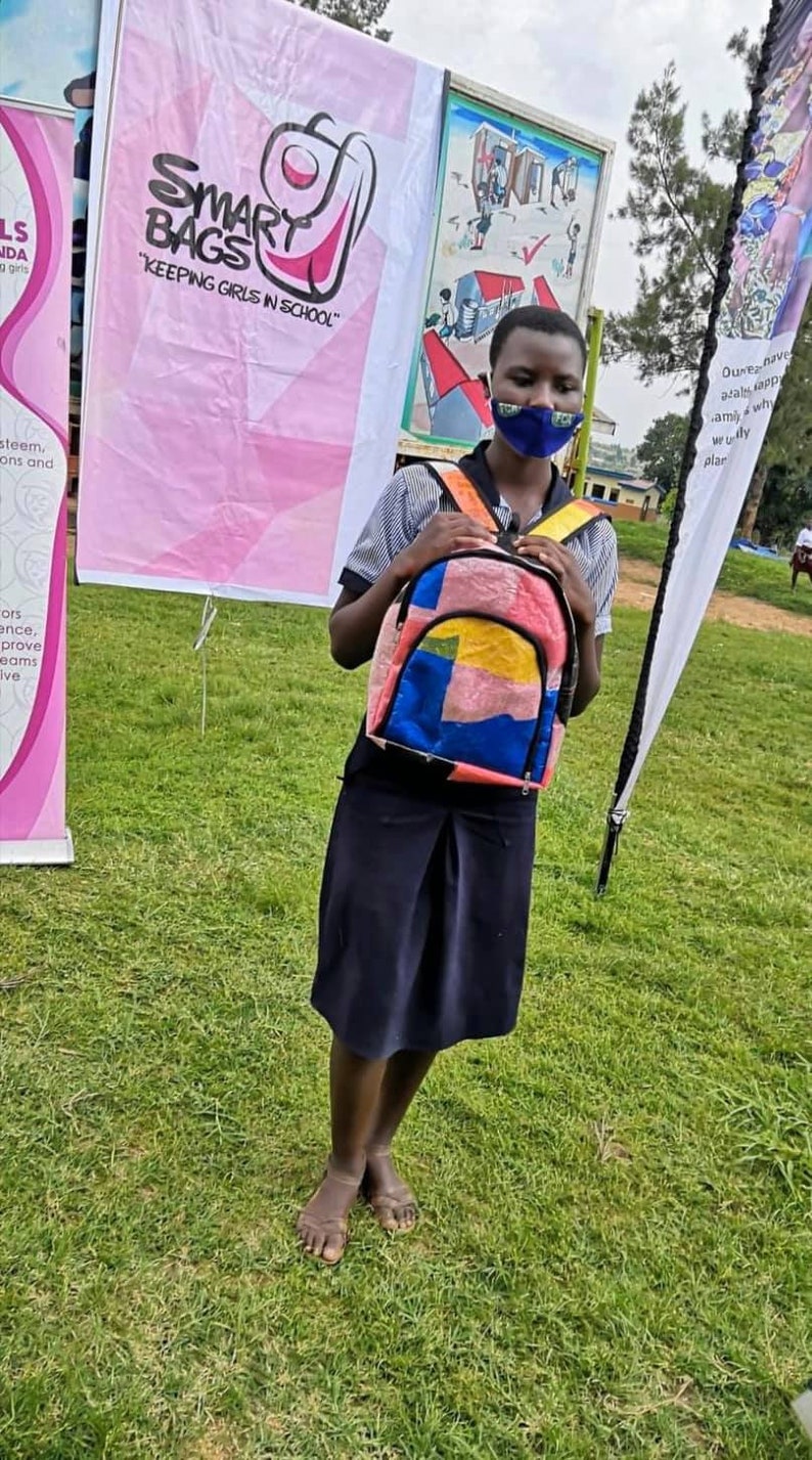 Pu&ograve; includere: Una giovane persona con una maschera facciale tiene uno zaino colorato con motivi multipli. Lo zaino ha una cerniera e spalline nere. La persona &egrave; in piedi su un prato erboso. Un cartello dietro la persona recita "SMART BAGS "KEEPING GIRLS IN SCHOOL."