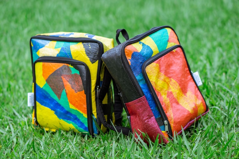 Pu&ograve; includere: Due zaini colorati realizzati con sacchetti di plastica riciclati. Gli zaini hanno spalline e cerniere nere. Gli zaini sono seduti su un letto di erba verde.