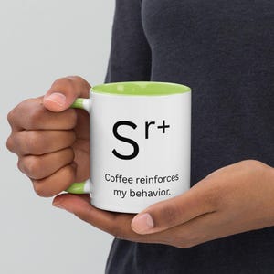Könnte beinhalten: Weißer Keramikbecher mit grünem Innenraum und Henkel. Der Becher zeigt den Text "Sr+" über dem Satz "Coffee reinforces my behavior." Ein humorvoller und stilvoller Becher für Kaffeeliebhaber.
