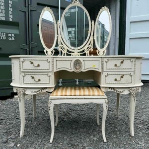 May include: A cream-coloured antique vanity set with a tri-fold mirror, drawers, and a matching stool. The vanity has gold accents and ornate detailing. The stool has a striped seat. A classic piece of bedroom furniture.