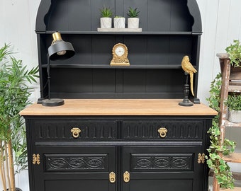 May include: Black and natural wood hutch with an arched top. The hutch features a black base with two doors and two drawers, adorned with gold hardware. The top section has shelves and a row of pegs. Decorative items include a clock, lamp, and bird.