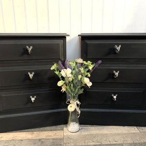 May include: Two black bedside tables, each with three drawers and silver deer head handles. A vase of flowers is placed between them. The tables are painted black and have a rectangular design.