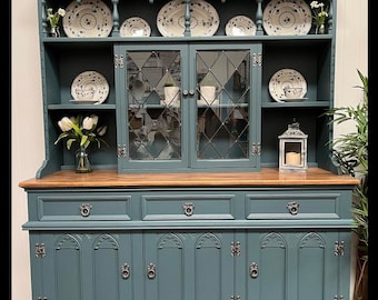 May include: A teal-colored hutch with a wooden top and base. The hutch features glass-paned doors, shelves displaying decorative plates, and ornate hardware. A candle lantern and vases with flowers add to the decor.