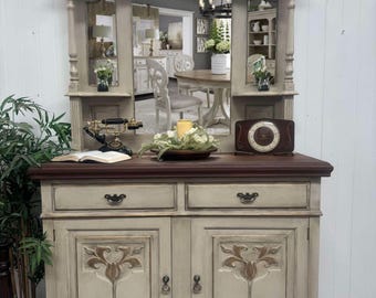 May include: A vintage-style, gray painted, wooden hutch with a mirror back and two doors. The hutch has a carved design on the top and bottom, and a brown wooden top.