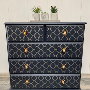 May include: A navy blue chest of drawers featuring a gold quatrefoil design on the drawer fronts. It has gold bunny-shaped handles and three small potted plants. A stylish storage unit for a bedroom or living space.