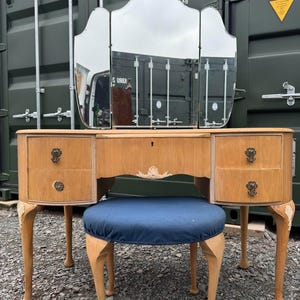 May include: A vintage wooden dressing table set with a three-panel mirror, two drawers, and a matching stool. The dressing table is light wood with brass hardware and the stool has a blue upholstered seat.