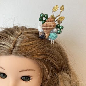 May include: A doll with blonde hair wearing a seashell hair accessory. The hair accessory features a brown seashell, green beads, gold glitter leaves, and a small turquoise seashell.