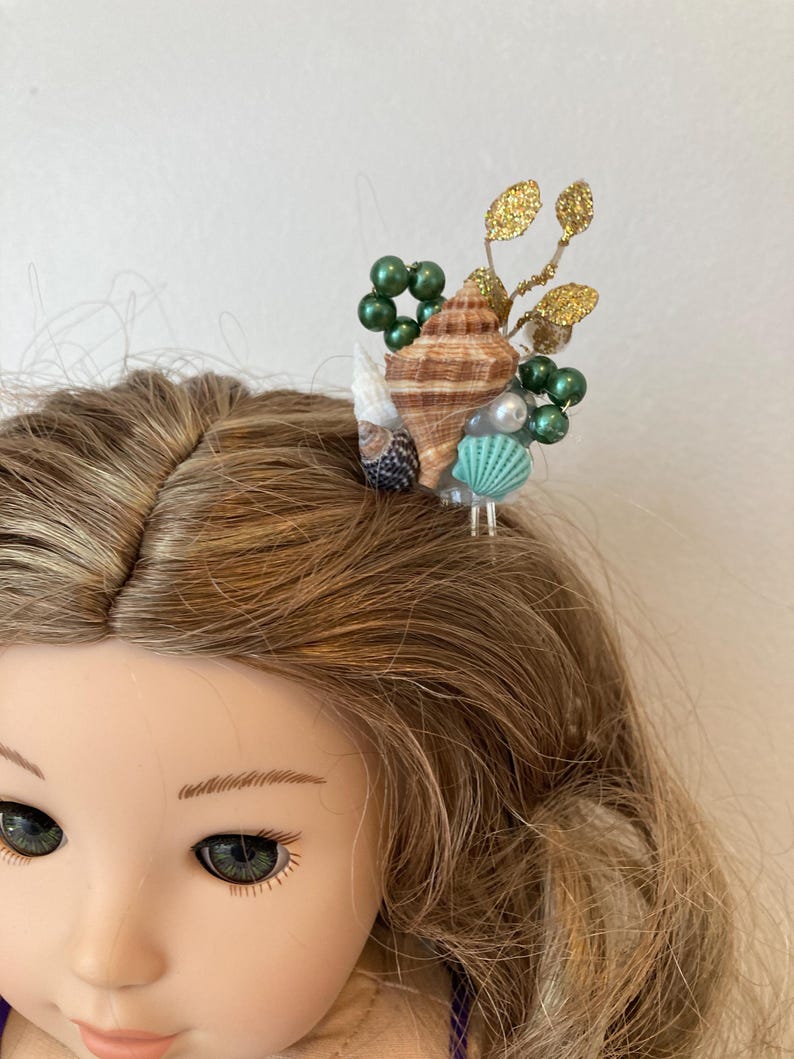 May include: A doll with blonde hair wearing a seashell hair accessory. The hair accessory features a brown seashell, green beads, gold glitter leaves, and a small turquoise seashell.