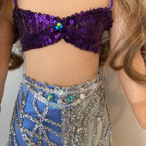 May include: A doll wearing a purple sequined top with a jewel accent and a two-tone skirt. The skirt is blue and silver with a net overlay. The doll's outfit is accessorized with a beaded belt.