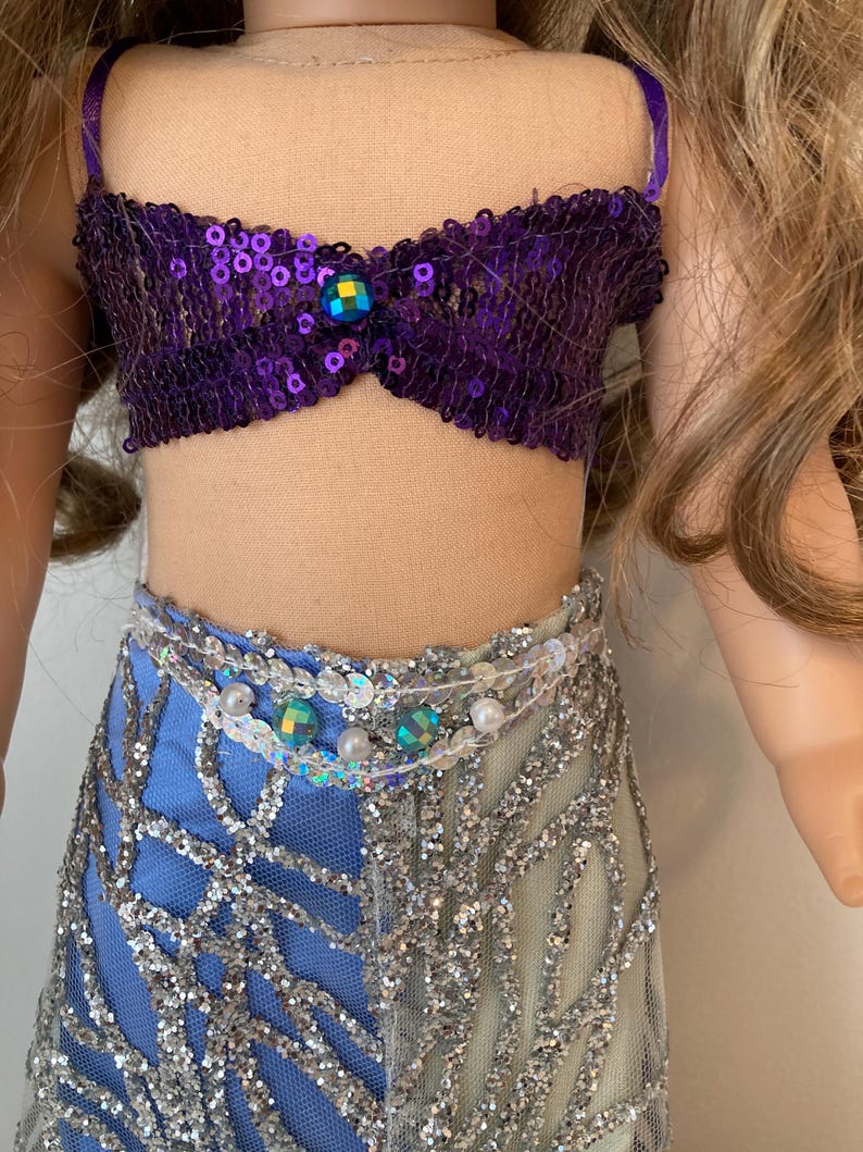 May include: A doll wearing a purple sequined top with a jewel accent and a two-tone skirt. The skirt is blue and silver with a net overlay. The doll's outfit is accessorized with a beaded belt.
