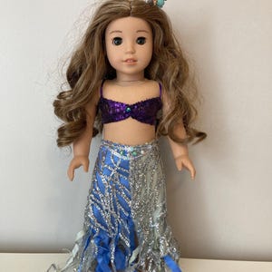 May include: A doll dressed as a mermaid, featuring a purple sequined top and a blue and silver skirt with flowing fabric. The doll has long, wavy brown hair and a seashell and bead hair accessory.