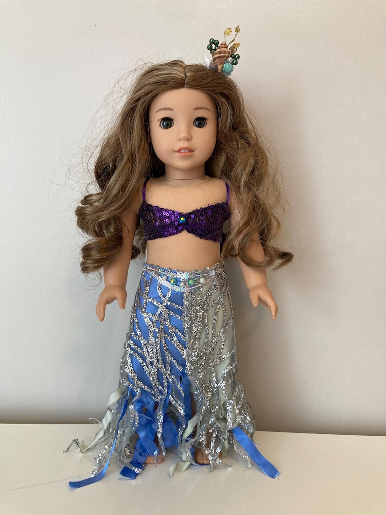 May include: A doll dressed as a mermaid, featuring a purple sequined top and a blue and silver skirt with flowing fabric. The doll has long, wavy brown hair and a seashell and bead hair accessory.