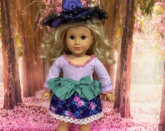 May include: A doll with blonde hair wearing a purple witch hat with black lace trim, a long-sleeved lavender top with ruffled edges, a sage green fabric bow at the waist, and a dark blue skirt featuring a pink floral pattern and white lace trim.