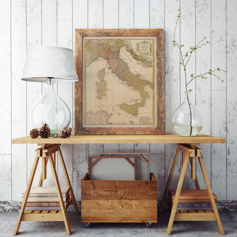Puede incluir: Un mapa vintage de Italia en un marco de madera cuelga sobre una mesa de madera con dos caballetes. La mesa est&aacute; decorada con un jarr&oacute;n de vidrio con una rama de hojas, una l&aacute;mpara de vidrio con una pantalla blanca y una pila de libros.