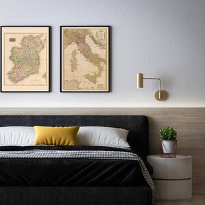Puede incluir: Dos mapas vintage enmarcados de Irlanda e Italia colgados en una pared blanca sobre una cabecera negra con una almohada amarilla y una colcha blanca. Una peque&ntilde;a planta en maceta se encuentra en una mesita de noche blanca junto a la cama.