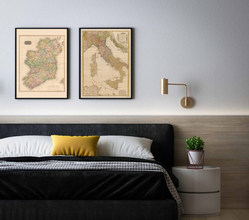 Puede incluir: Dos mapas vintage enmarcados de Irlanda e Italia colgados en una pared blanca sobre una cabecera negra con una almohada amarilla y una colcha blanca. Una peque&ntilde;a planta en maceta se encuentra en una mesita de noche blanca junto a la cama.