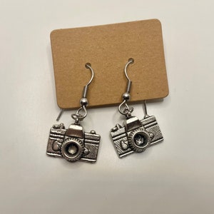 Puede incluir: Un par de pendientes de cámara plateados con un dije de cámara colgante. Los pendientes están en una tarjeta marrón.