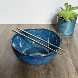 May include: A deep blue ceramic bowl with an irregular, organic rim and a speckled glaze finish. Two slender, polished silver-toned metal chopsticks rest diagonally across the top of the bowl, positioned within the notched edges of the rim.