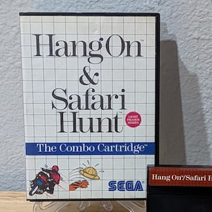 Puede incluir: Un cartucho combinado de videojuegos SEGA retro, "Hang On & Safari Hunt", con su caja y manual originales. La caja presenta un patrón de cuadrícula e ilustraciones de una motocicleta y un sombrero de safari. El cartucho del juego es negro con texto rojo.