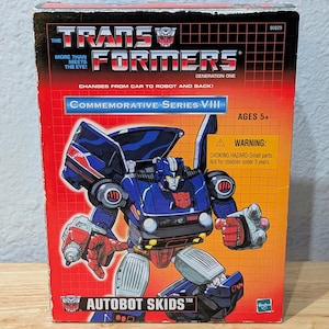 May include: A vintage Transformers Generation One Autobot Skids toy box. The box is red and orange with a blue and red robot illustration. Text includes "More Than Meets The Eye!" and "Commemorative Series VIII". Ages 5+ warning is also present.