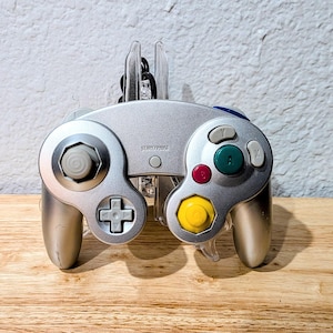 Pode incluir: Controlador de videogame prateado com botões coloridos, incluindo verde, vermelho e amarelo. O controlador tem um direcional, um joystick e um botão iniciar/pausar. O controlador está sobre uma superfície de madeira.