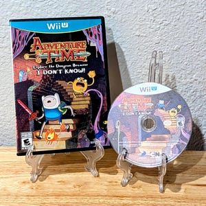 May include: A Wii U video game case and disc for "Adventure Time: Explore the Dungeon Because I DON'T KNOW!" The case features cartoon characters and the game title. The disc is displayed next to the case on a clear stand.