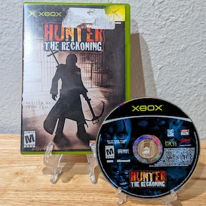 Puede incluir: Caja y disco del juego de Xbox "Hunter: The Reckoning". La caja es verde con el título del juego en rojo y blanco, y una figura silueteada sosteniendo una ballesta. El disco presenta el título del juego y un diseño oscuro y ominoso.