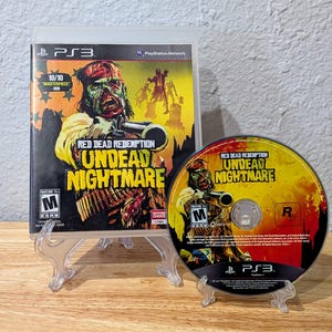 May include: A PlayStation 3 game case and disc for "Red Dead Redemption: Undead Nightmare." The cover art features a zombie holding a gun, with the game title in yellow. The disc has similar artwork. The game is rated M for Mature.