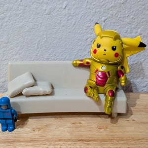Pu&ograve; includere: Una figura gialla di Pikachu in un'armatura dorata e rossa in stile Iron Man &egrave; seduta su un piccolo divano bianco con due cuscini bianchi. Una action figure blu &egrave; vicina. Le figure sono su una superficie di legno.