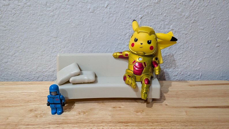 Pu&ograve; includere: Una figura gialla di Pikachu in un'armatura dorata e rossa in stile Iron Man &egrave; seduta su un piccolo divano bianco con due cuscini bianchi. Una action figure blu &egrave; vicina. Le figure sono su una superficie di legno.