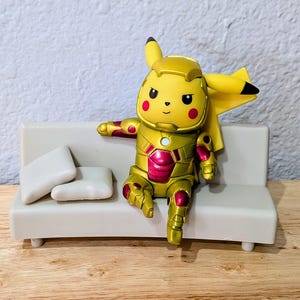 Pu&ograve; includere: Una figura gialla di Pikachu con un'armatura dorata e rossa, seduta su un piccolo divano bianco con due cuscini bianchi. La figura ha occhi neri e guance rosse. Il divano &egrave; su una superficie di legno.