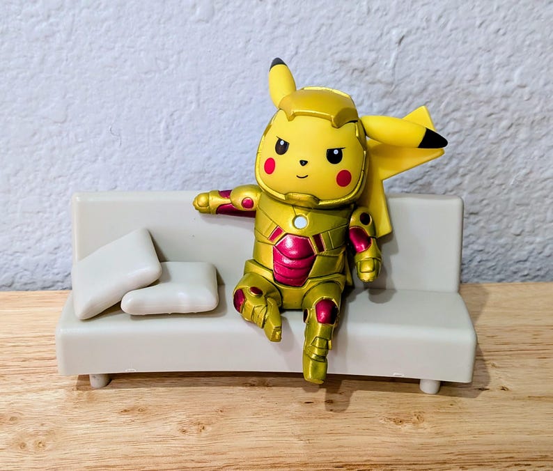 Pu&ograve; includere: Una figura gialla di Pikachu con un'armatura dorata e rossa, seduta su un piccolo divano bianco con due cuscini bianchi. La figura ha occhi neri e guance rosse. Il divano &egrave; su una superficie di legno.
