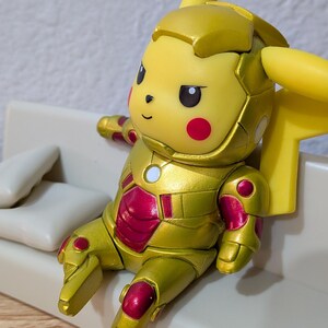 Pu&ograve; includere: Una figura gialla di Pikachu in un'armatura dorata e rossa, seduta su un piccolo divano bianco sporco con due cuscini. La figura ha orecchie con la punta nera e guance rosse. Il divano &egrave; su una superficie di legno.
