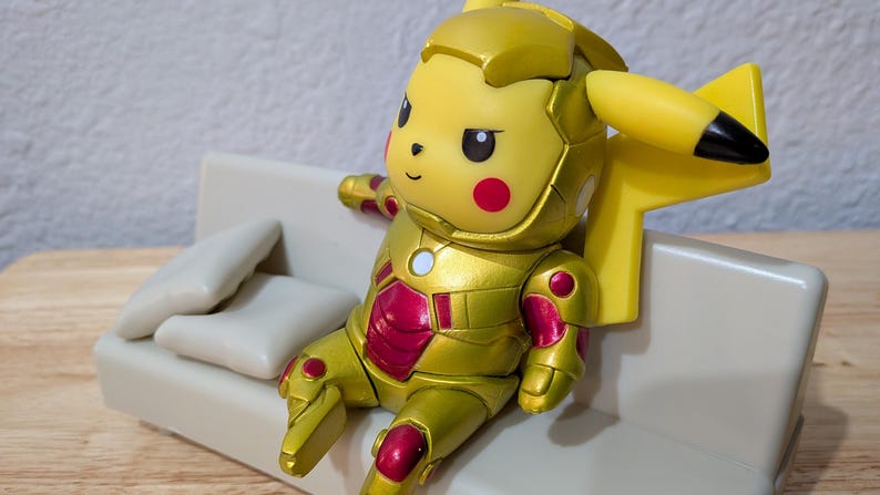 Pu&ograve; includere: Una figura gialla di Pikachu in un'armatura dorata e rossa, seduta su un piccolo divano bianco sporco con due cuscini. La figura ha orecchie con la punta nera e guance rosse. Il divano &egrave; su una superficie di legno.