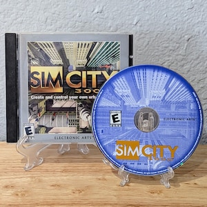 May include: A SimCity 3000 PC game in its original case. The game case is silver with the game title in gold and the text "Create and control your own urb". The CD is blue with the game title in gold.