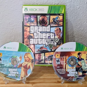 May include: An Xbox 360 video game case and two game discs for Grand Theft Auto V. The case is green and features a collage of characters and scenes. The discs have colourful graphics and the game title.
