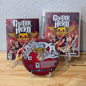 Op de afbeelding: Een Guitar Hero Aerosmith videogame voor PlayStation 3. De gamecase en disc tonen het logo van de game en de bandleden van Aerosmith. De disc is rood met witte tekst en afbeeldingen. De gamecase is rood en zwart.