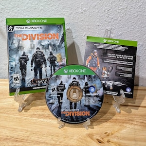 以下が含まれることがあります： 「Tom Clancy's The Division」のXbox Oneビデオゲームケースとディスク。ゲームケースは緑色で、ゲームタイトルとキャラクターが前景にあります。ディスクも同じ画像です。プロモーションインサートも表示されています。