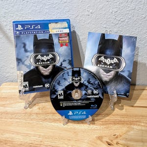 Puede incluir: Un juego de video PS4 que presenta "Batman: Arkham VR". La caja del juego, el disco y la inserción presentan la imagen de Batman. El juego está clasificado como "M" para audiencias maduras. El juego se muestra sobre una superficie de madera.