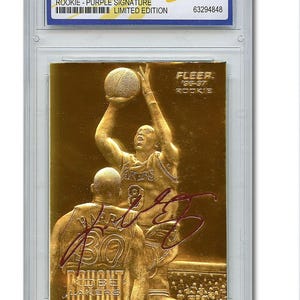 May include: A gold-colored basketball card featuring Kobe Bryant in action, with a purple signature. The card is from the 1996-97 Fleer 23KT Gold series, labeled as a rookie card, and is graded WCG 10 GEM-MT.