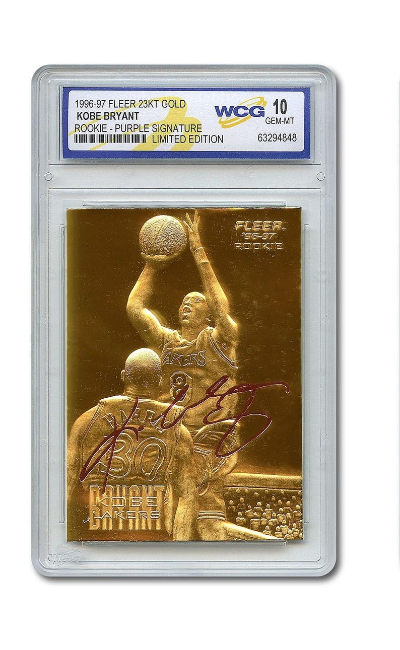 May include: A gold-colored basketball card featuring Kobe Bryant in action, with a purple signature. The card is from the 1996-97 Fleer 23KT Gold series, labeled as a rookie card, and is graded WCG 10 GEM-MT.