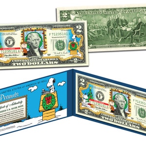 May include: A two dollar bill featuring a Peanuts Christmas theme. The bill is colored with Snoopy typing on a typewriter and Charlie Brown standing next to a Christmas tree. The bill is framed in a blue border and includes a certificate of authenticity.