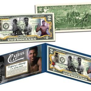 May include: A two-dollar bill featuring portraits of Cassius Clay, with a gold-colored frame. The bill includes the text "United States of America" and "Two Dollars". A certificate of authenticity is also visible.