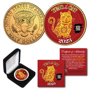 May include: A gold-colored coin with a red background featuring a stylized image of a cat with the text "Year of the Cat" and "2023" in gold. The coin is in a black velvet box with a certificate of authenticity.