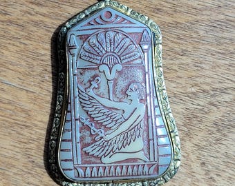 May include: A gold-toned pendant with an intricate design featuring an Egyptian goddess with wings, a sun disc, and an ankh symbol. The pendant is made of metal and has a red and white color scheme.