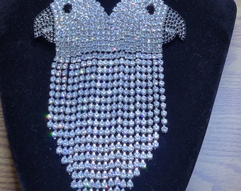 May include: A dazzling, silver-toned brooch shaped like two stylized birds, encrusted with sparkling rhinestones. The birds' bodies transition into a cascade of dangling rhinestone strands, creating a glamorous, eye-catching accessory. The brooch is displayed on a black velvet surface.