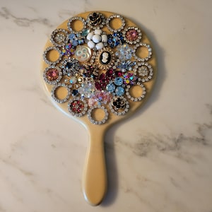 May include: A vintage hand mirror with a cream-colored handle and a round mirror face. The mirror face is completely covered with a mosaic of rhinestones, jewels, and buttons in various colors, including red, blue, pink, and white.