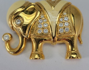 May include: Gold-toned elephant brooch with cream-colored enamel accents. The elephant is adorned with clear, sparkling rhinestones and features intricate detailing. A vintage-style jewelry piece.